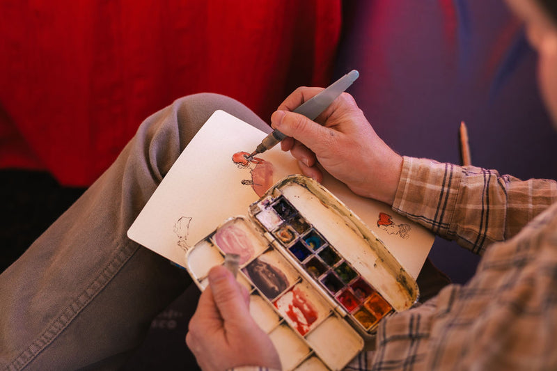 Hands sketching with a pen over a pocket watercolor palette