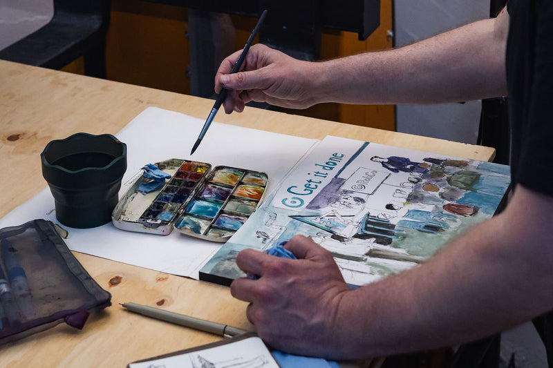Artist painting in a sketchbook beside a watercolor palette